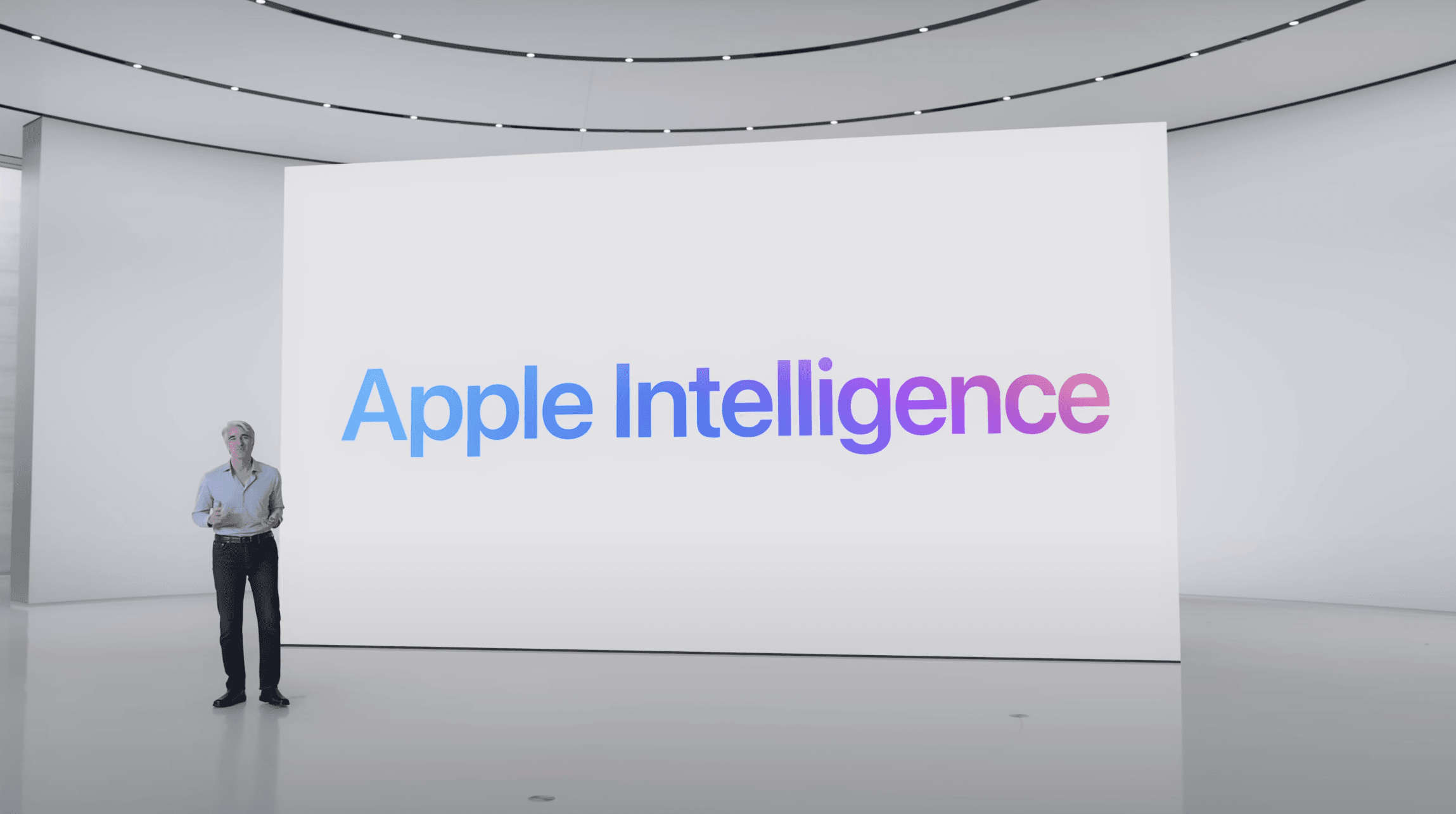 Craig Federighi standing on stage in a modern, minimalist auditorium, giving a presentation during the keynote of WWDC 2024. Behind him is a large screen displaying the text 'Apple Intelligence' in gradient blue and purple colors.